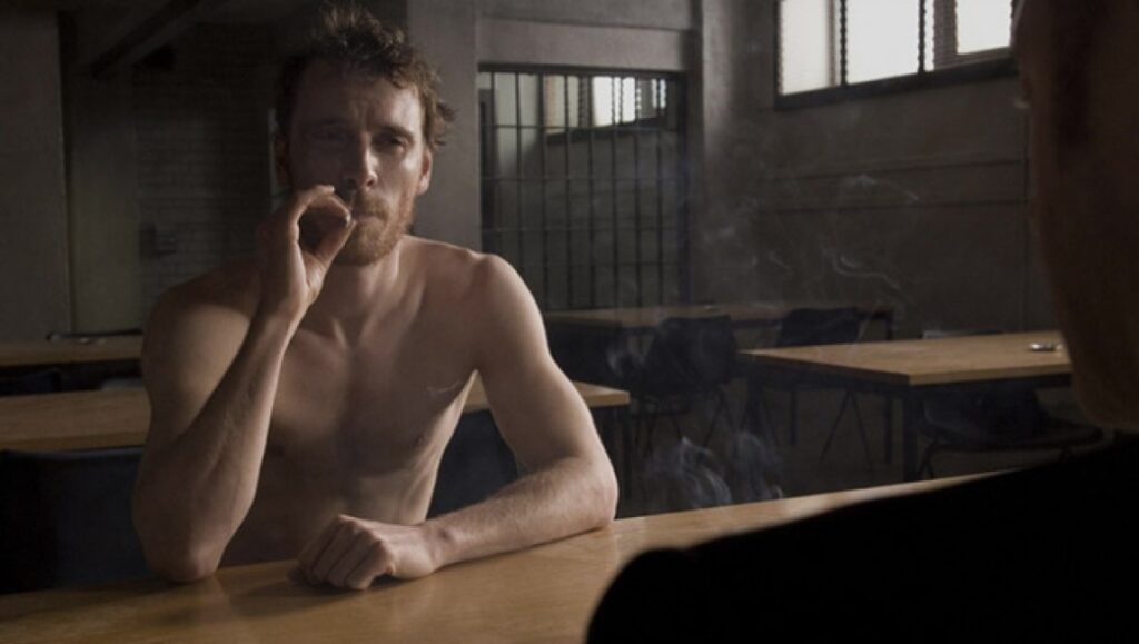 Michael Fassbender in Hunger movie. Bare-chested man smoking in a prison cell. Steve McQueen film.