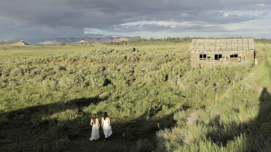 Butter on the Latch film still. Two women walk toward an old cabin in a field under a stormy sky. Josephine Decker film.