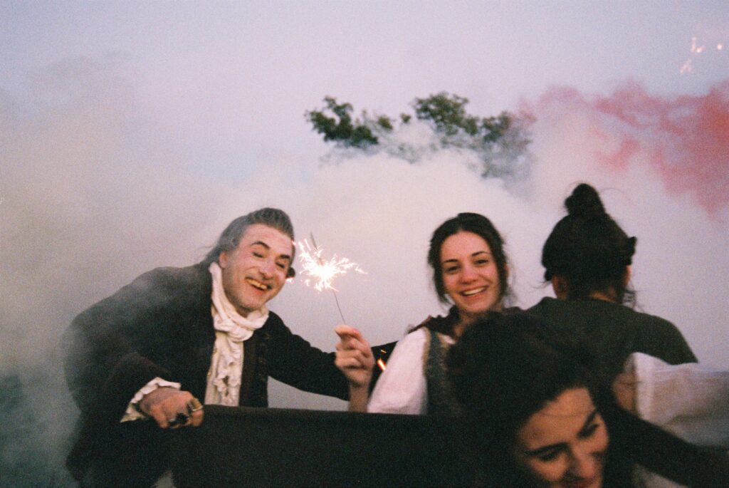 Story of My Death film still. People in period clothing smile amidst smoke, one holding a sparkler. Albert Serra film.