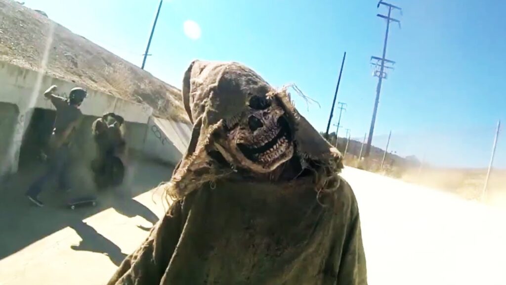 V/H/S: Viral horror film still. A creepy, hooded skull mask figure faces the camera. Skateboarders in the background.