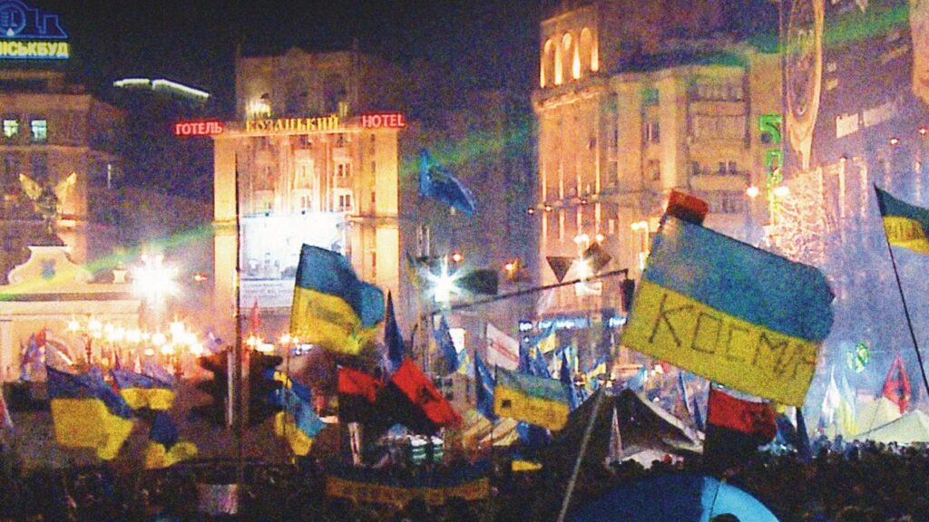 Maidan | Sergei Loznitsa Maidan Square scene. Ukrainian flags wave amidst crowds, buildings lit up at night. Sergei Loznitsa film.