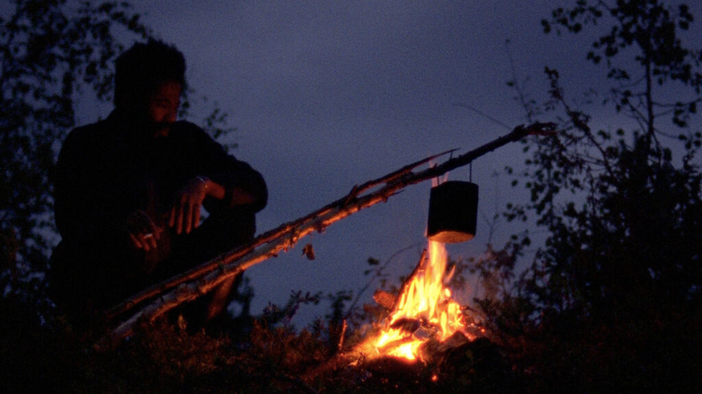 A Spell to Ward Off the Darkness | Ben Rivers & Ben Russell A Spell to Ward Off the Darkness: Dark silhouette of a person by a campfire, cooking pot hanging above flames. Night scene.