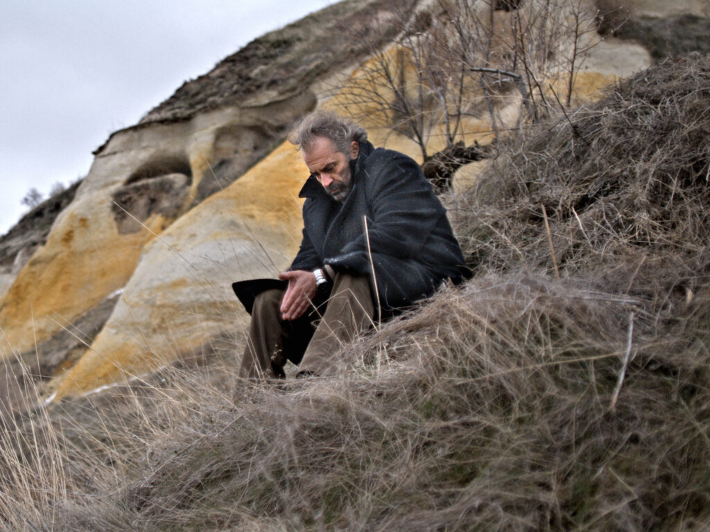 Nuri Bilge Ceylan's Winter Sleep still: Man sits pensively on a hill. Winter landscape with dry grass and rock formations.