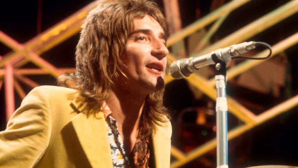Rod Stewart performing Every Picture Tells a Story. Singer with microphone in yellow jacket.