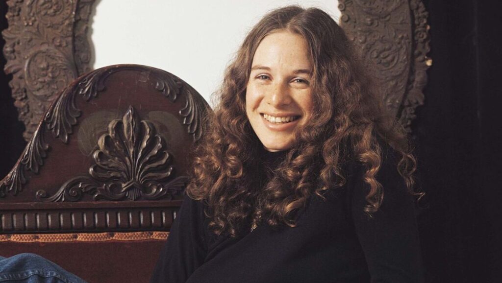 Carole King, Tapestry era. Singer-songwriter with curly brown hair smiling, seated in an ornate chair.