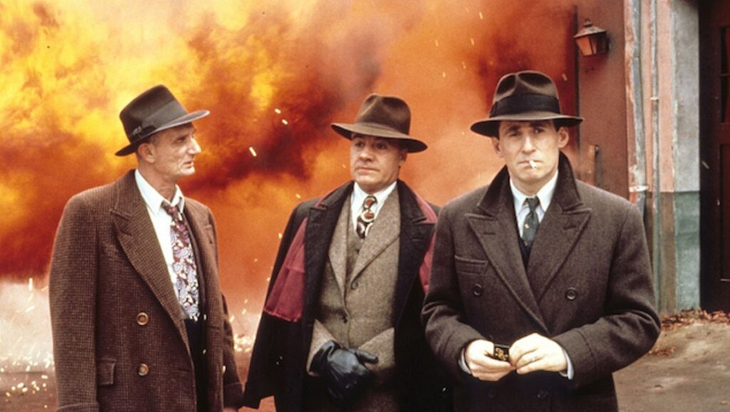 Miller’s Crossing | Joel & Ethan Coen Miller's Crossing movie still. Three men in hats and coats stand before an explosion. Coen Brothers film.