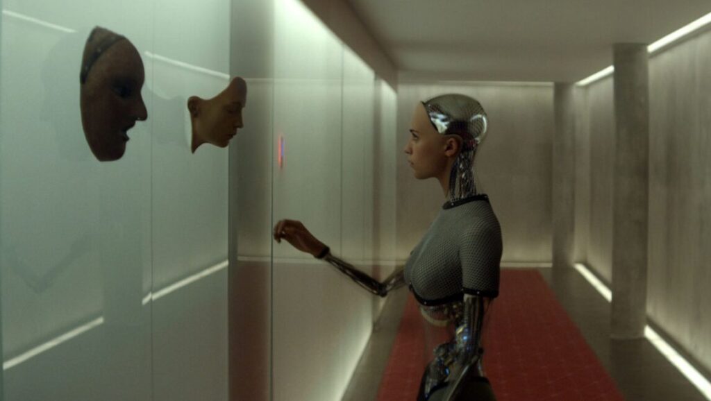 Ex Machina Review | Alex Garland Ex Machina movie scene. Ava, a humanoid robot, interacts with a wall display in a modern corridor.