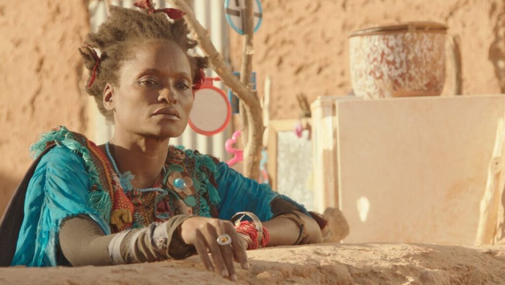 Timbuktu | Abderrahmane Sissako Abderrahmane Sissako's Timbuktu film still. Woman in colorful clothing, jewelry, and unique hairstyle. Desert backdrop.