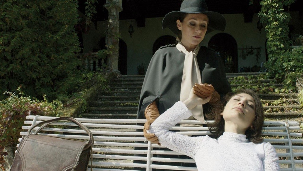 The Duke of Burgundy film still. Woman in hat and cape with woman reclining on bench.
