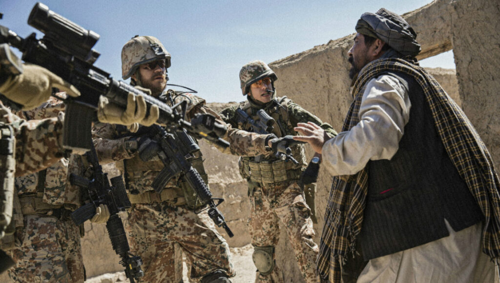 Scene from A War movie. Soldiers with guns confront a local man in a war-torn area.