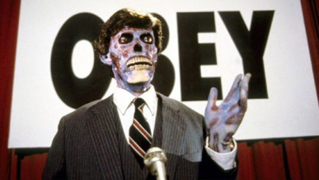 They Live movie still. Alien in disguise, wearing a suit, stands before an OBEY sign. John Carpenter film.