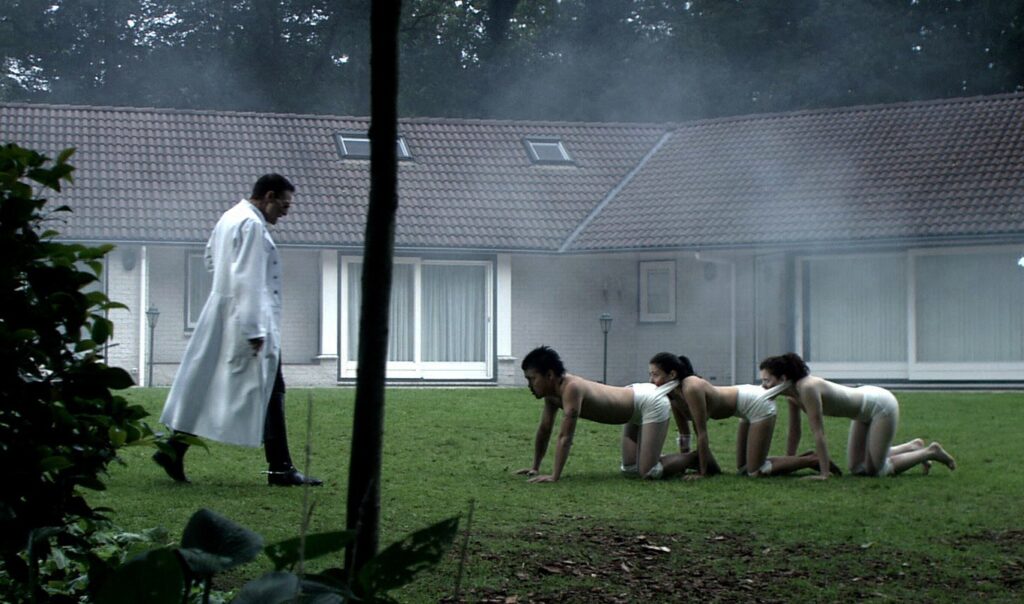 The Human Centipede (First Sequence) | Tom Six The Human Centipede movie still. Doctor watching three people on all fours, mouths sewn to the next person's anus.