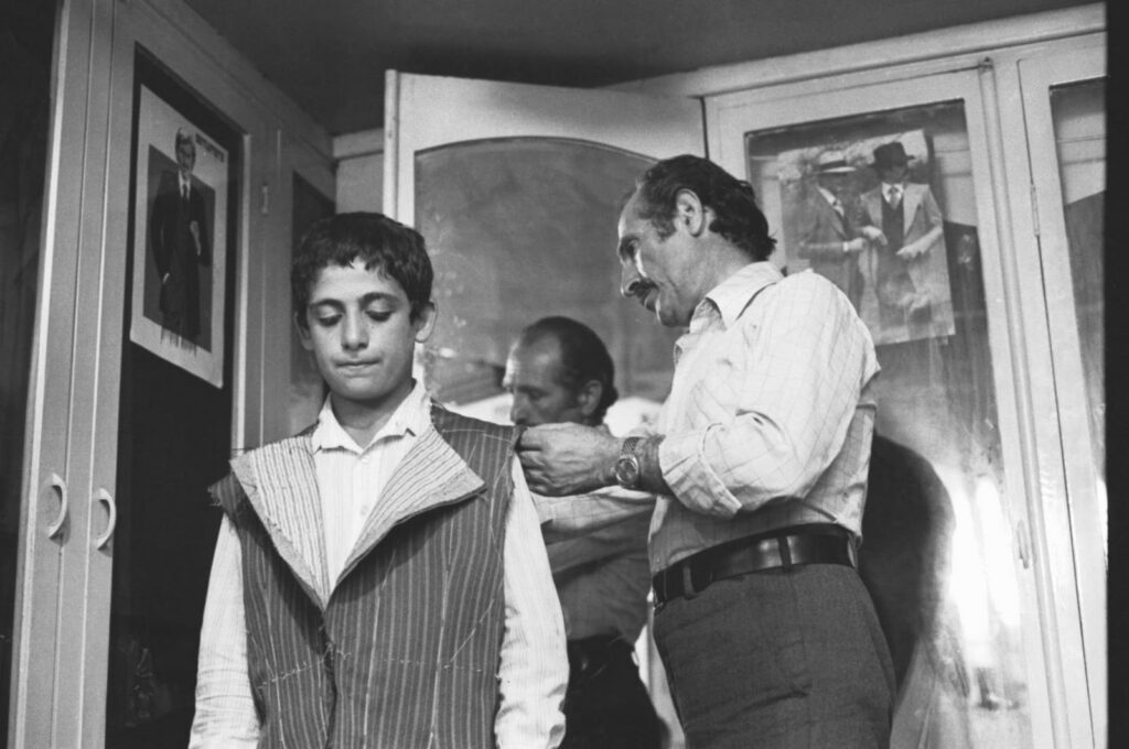 Black and white film still from A Suit for Wedding by Abbas Kiarostami. Tailor fitting a striped suit vest on a young man.
