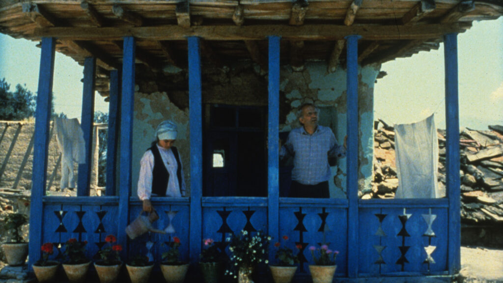 Life, and Nothing More… | Abbas Kiarostami Life, and Nothing More... film still. Woman waters flowers on a blue porch, man stands behind. Directed by Abbas Kiarostami.