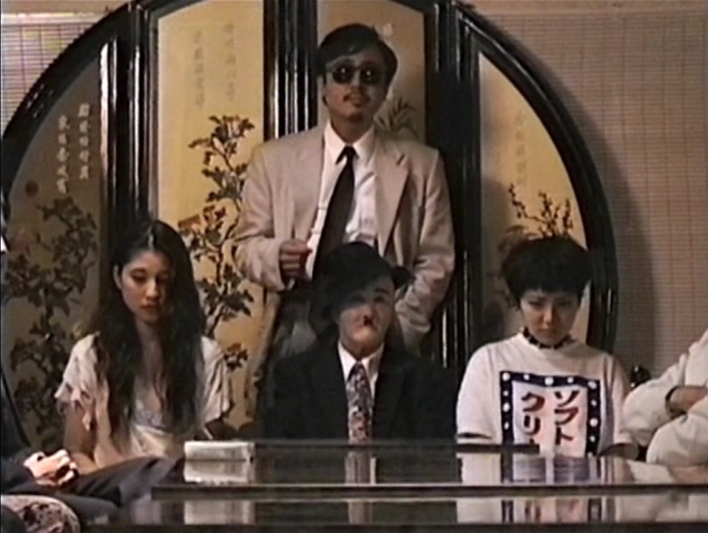 Bad Film scene by Sion Sono. Group of people seated at a table, including one in sunglasses and another in a hat.