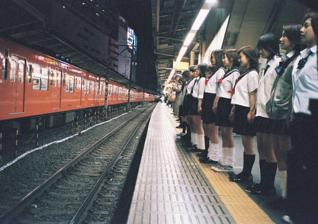 Suicide Club film still: Japanese schoolgirls line a train platform, awaiting a red train. Dark, eerie.