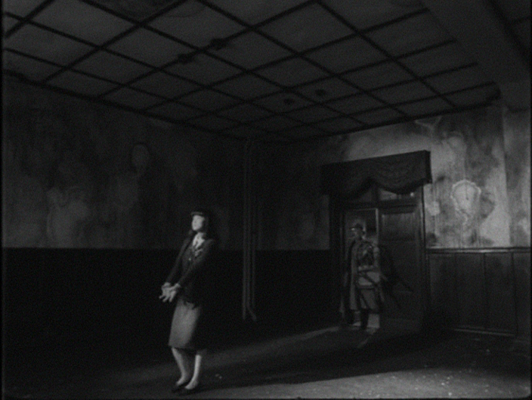 Black and white still from Sion Sono's The Room film. Woman in room, man in doorway.