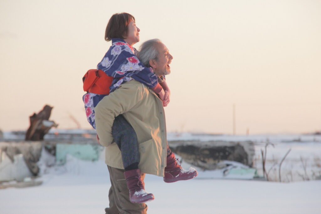 The Land of Hope film still: Elderly man gives a young woman a piggyback ride in a snowy landscape.