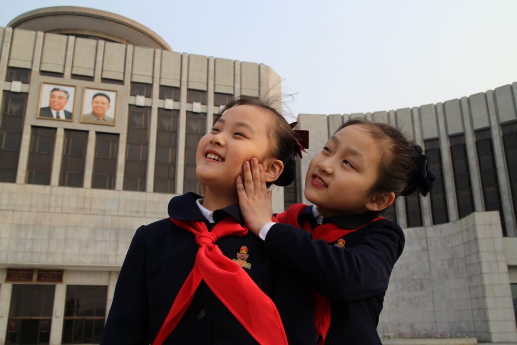 Under the Sun | Vitali Mansky Two girls in Under the Sun by Vitali Mansky. North Korean children smile, wearing red scarves, with portraits of leaders in background.