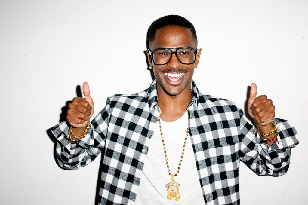 Big Sean gives thumbs up. Rapper promoting his album, I Decided. Wearing glasses, chain, and plaid shirt.