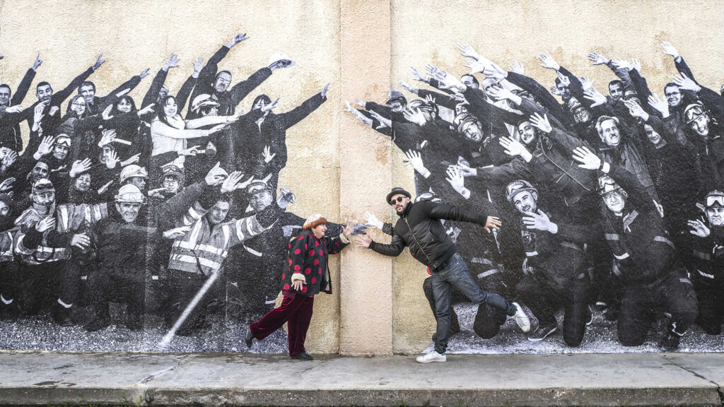 Faces Places at TIFF 2017: Agnès Varda and JR interact with a mural of people with raised arms. Film festival.