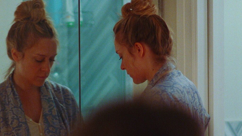 Golden Exits | Alex Ross Perry Golden Exits film still: Woman with bun looks in mirror, wearing patterned robe.