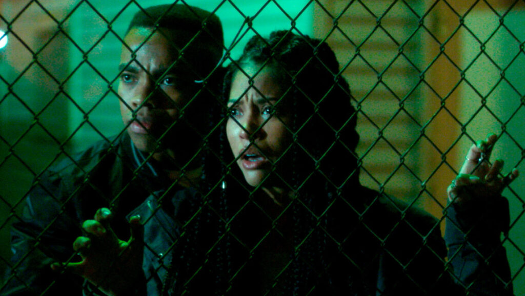 From The First Purge, a man and woman look scared behind a chain-link fence, lit with green light.