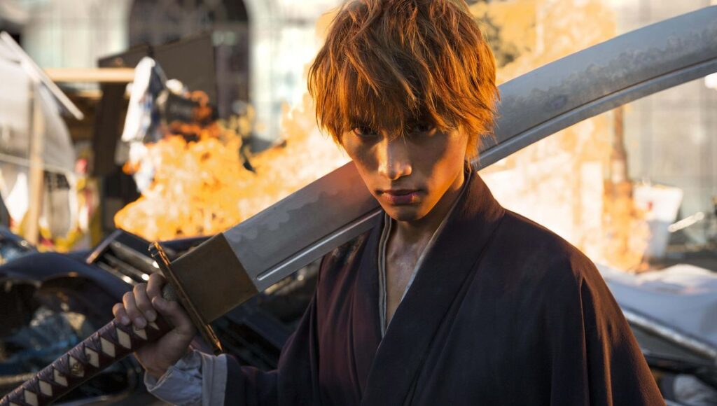 Ichigo Kurosaki in *Bleach*, directed by Shinsuke Sato. Ichigo holds a sword amidst a fiery, destroyed backdrop.