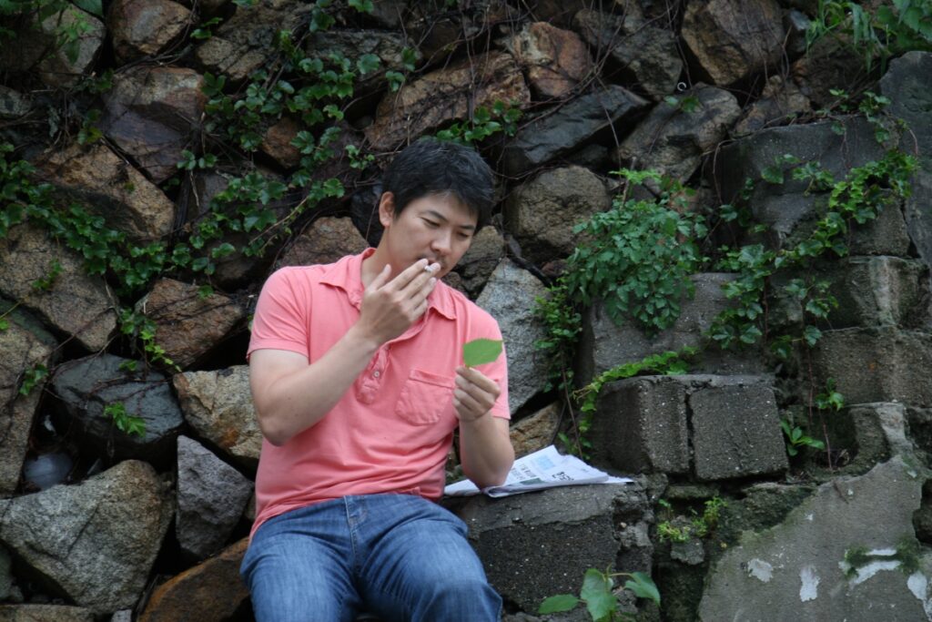 Hong Sang-soo's Hahaha film still. Man smokes, holds leaf. Stone wall backdrop. Korean cinema.