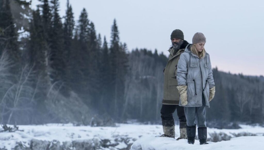 Hold the Dark movie scene. Man and woman in winter gear stand in snowy landscape. Dark forest backdrop.