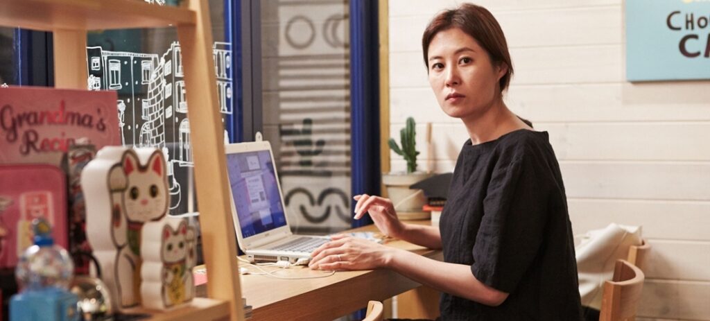 Hong Sang-soo Retrospective: Woman at laptop. Lucky cat statue, cactus, and Grandma's Recipe book visible.