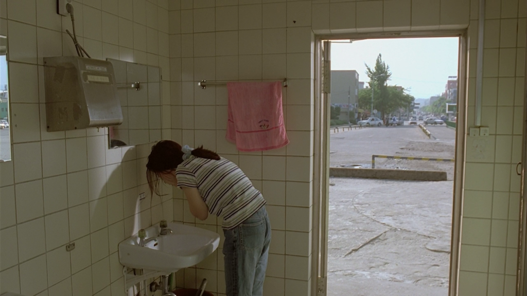 Scene from The Power of Kangwon Province: Woman washing face in tiled bathroom, street visible through doorway. Hong Sang-soo film.