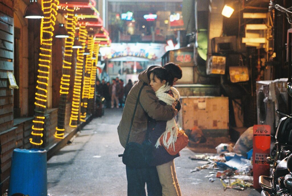Tale of Cinema | Hong Sang-soo Tale of Cinema film still. A couple embraces in a brightly lit alley, directed by Hong Sang-soo. Korean cinema.
