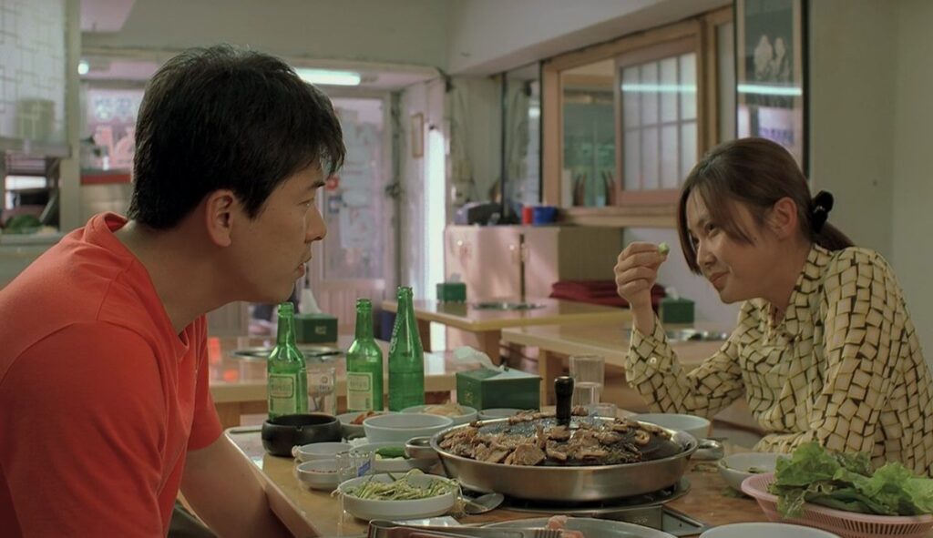 Turning Gate film still. Man and woman at Korean BBQ, soju bottles on table. Hong Sang-soo movie scene.