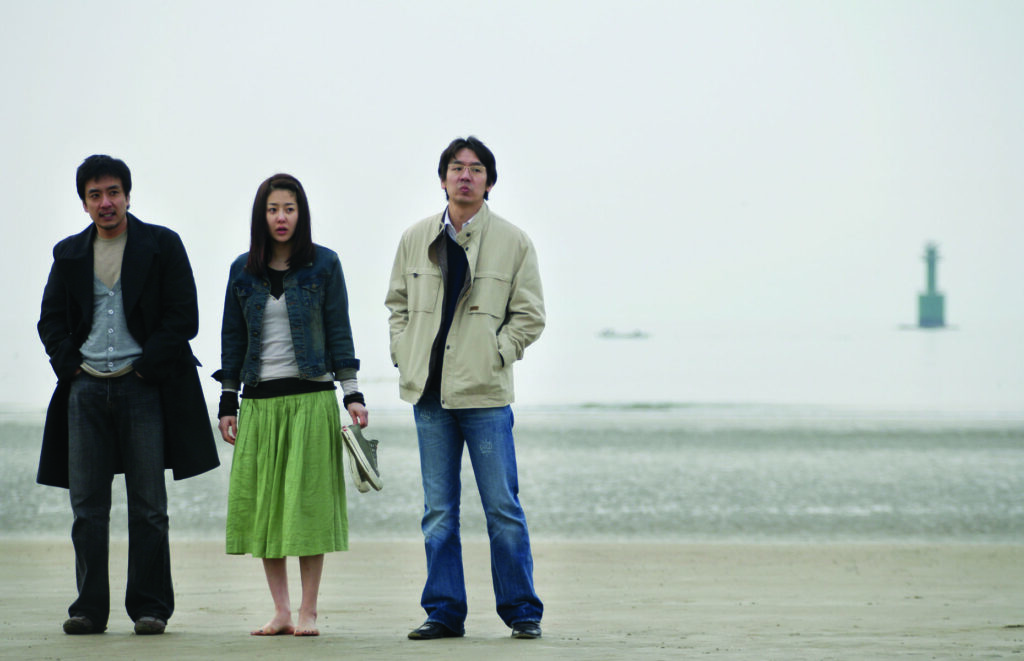 Woman on the Beach film still. Three people stand on a beach. Lighthouse in background.