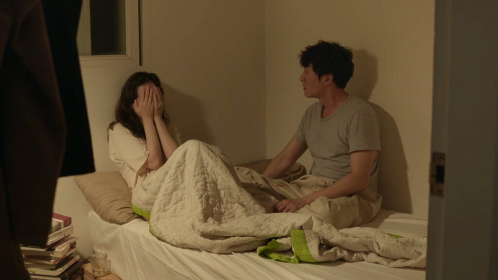 Yourself and Yours film scene. Woman covers face, man sits beside her on bed. Korean film, Hong Sang-soo movie.