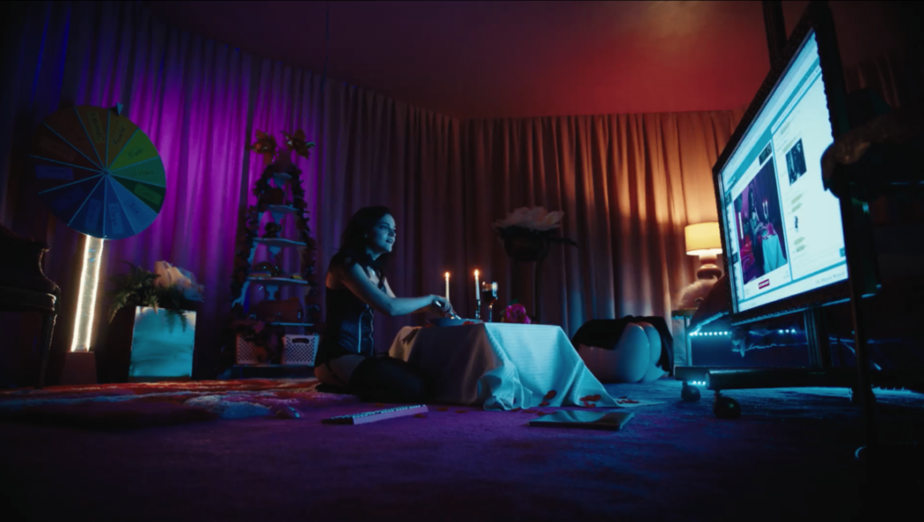 Cam movie scene: Woman at table with candles, large screen showing a cam feed in a colorful room.
