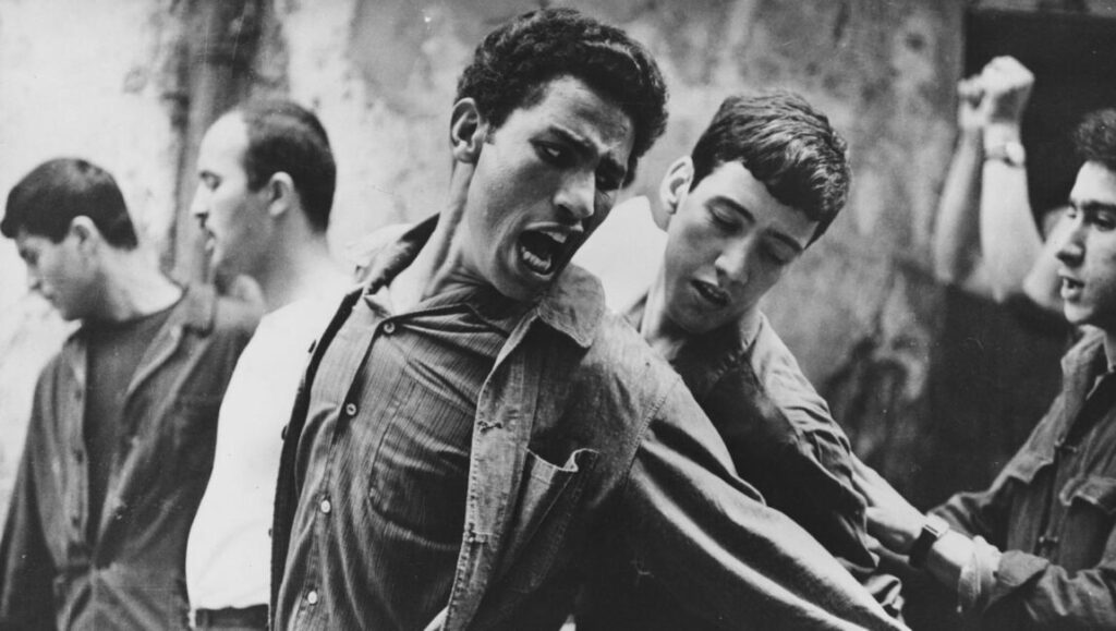 The Battle of Algiers film still. Black and white image shows a man shouting, held by others. Drama, conflict.