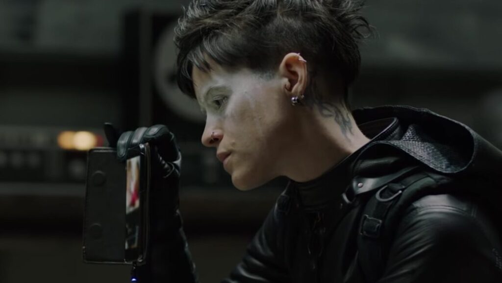 Claire Foy as Lisbeth Salander in The Girl in the Spider's Web movie. Foy holds a smartphone, wearing black leather.