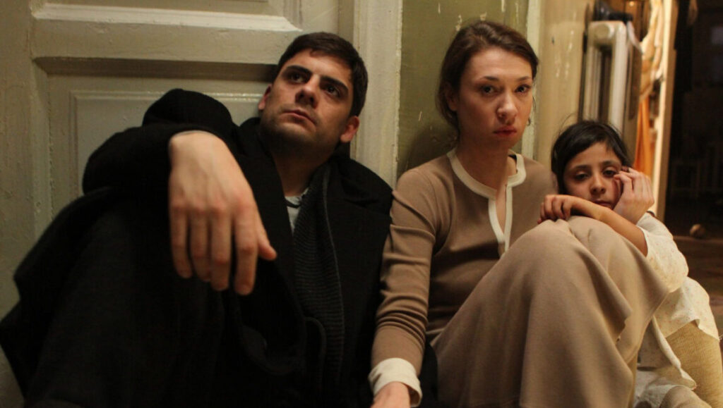 Dovlatov film still. A man, woman, and child sit against a wall. Alexey German Jr. film.