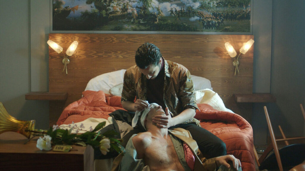 Neïl Beloufa's Occidental artwork. Two men on a bed, one tending to the other's head wound. Hotel room setting.