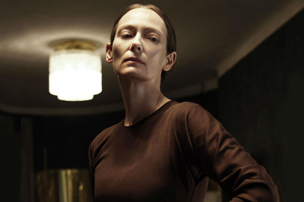 Tilda Swinton in Suspiria, a 2018 off-beat horror blockbuster movie. Dark, moody lighting.