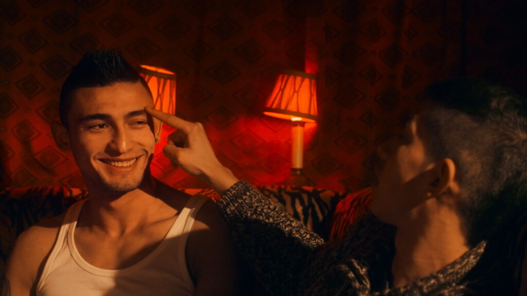 Brothers of the Night film still. Two young men, one smiling, in an intimate moment. Red lighting.