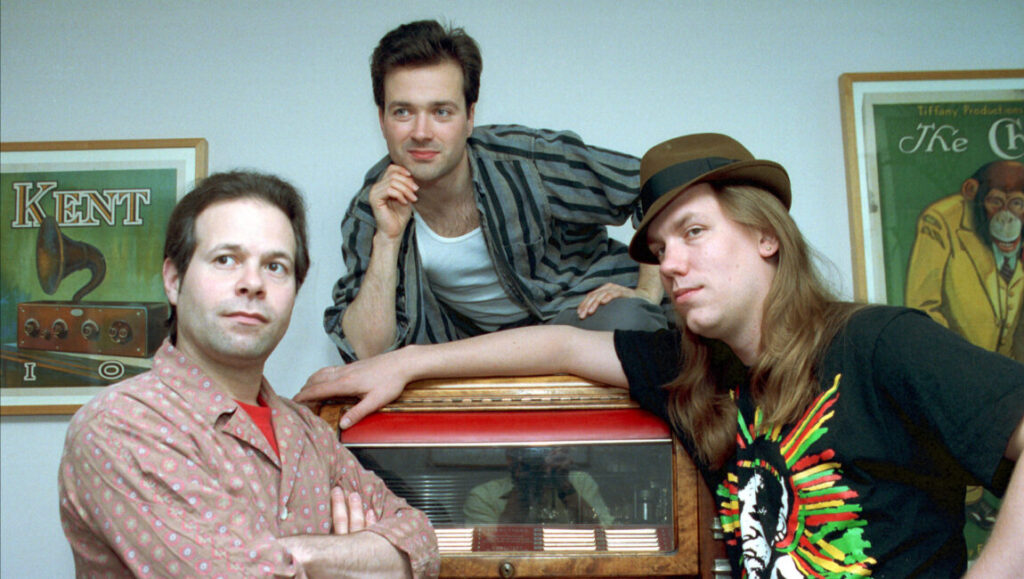 Violent Femmes | Violent Femmes Violent Femmes band portrait. Group members posing with vintage radio and posters. Alternative rock band image.