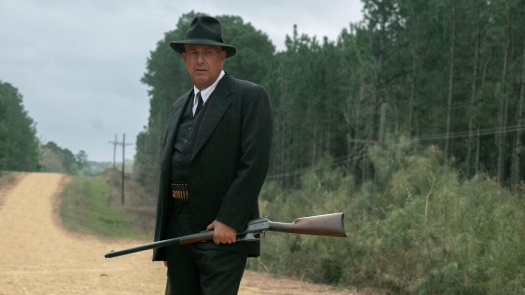 The Highwaymen | John Lee Hancock The Highwaymen movie still. Man in suit, hat, and gun belt holds rifle. John Lee Hancock film.