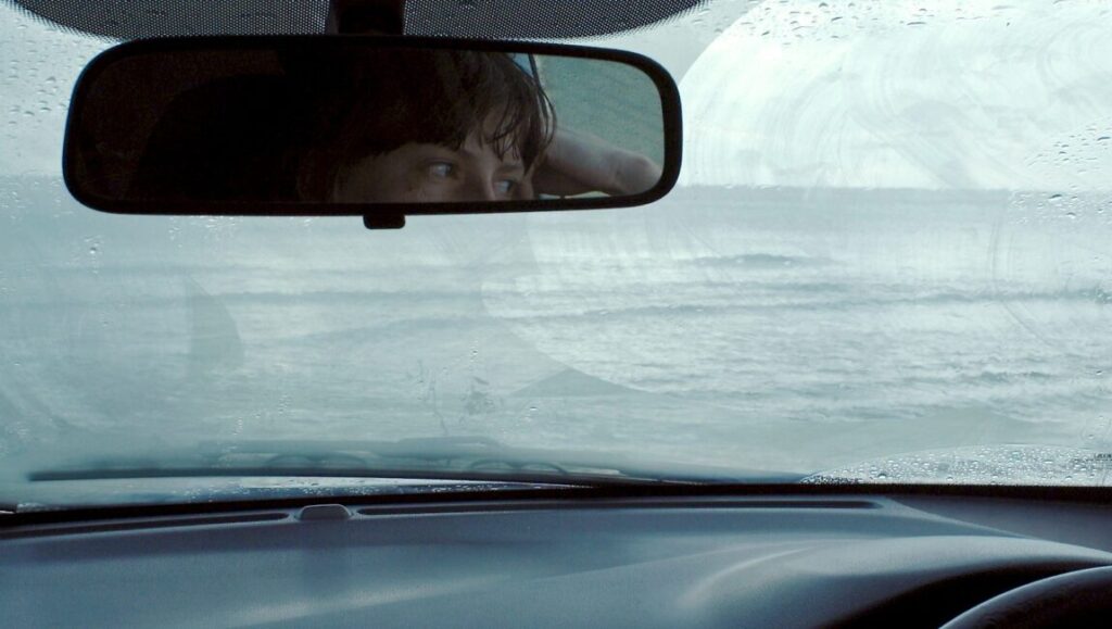 Drift | Helena Wittmann From inside a car, a view of the ocean is reflected in the rearview mirror in Drift by Helena Wittmann.