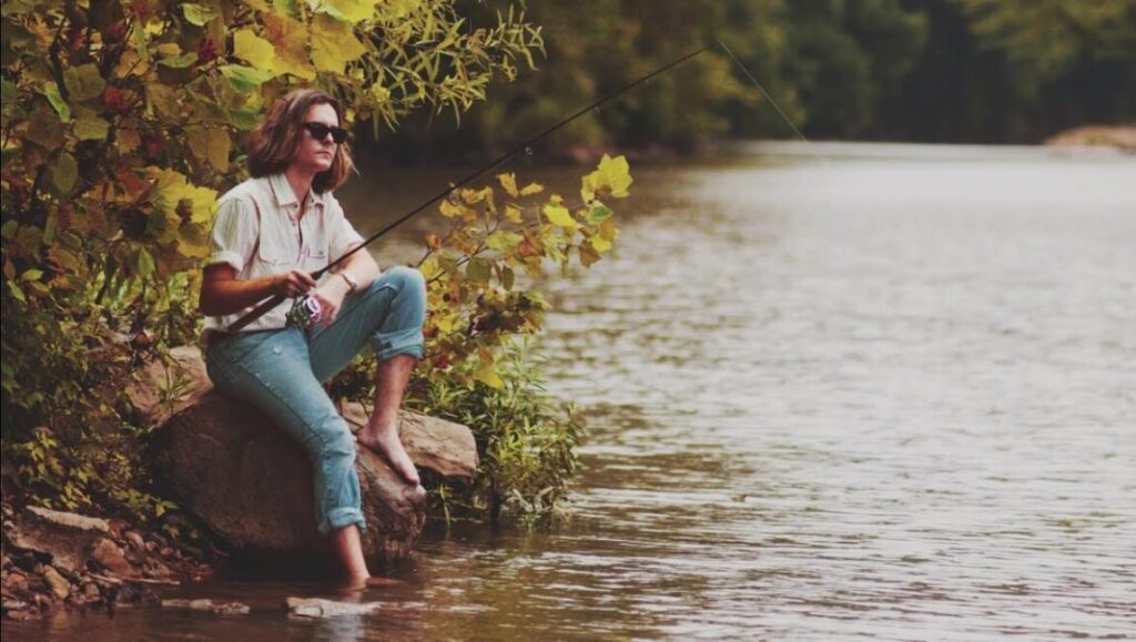 Rooted & Restless: Woman fishing. Angler with fishing rod sits by river. Outdoor recreation and relaxation.