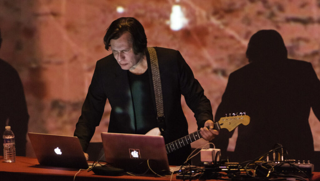 Foreign Correspondent | Issue 3 Foreign Correspondent: Musician with guitar, laptops, and audio equipment on stage. Live music performance.