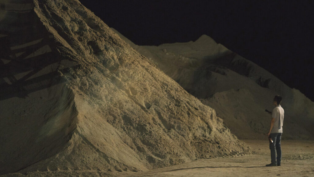 A Land Imagined film still. Man stands near sand piles at night. Yeo Siew Hua movie scene.