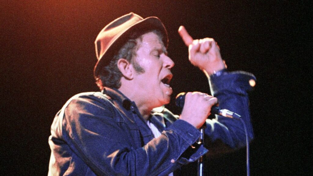 Tom Waits performing Mule Variations. Singer with hat, microphone, and expressive hand gesture. Concert stage lighting.